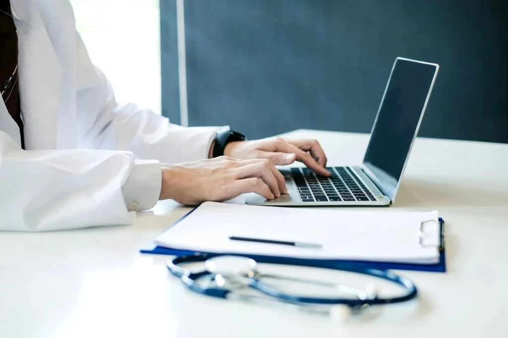 Physician typing on a laptop beside a stethoscope and clipboard